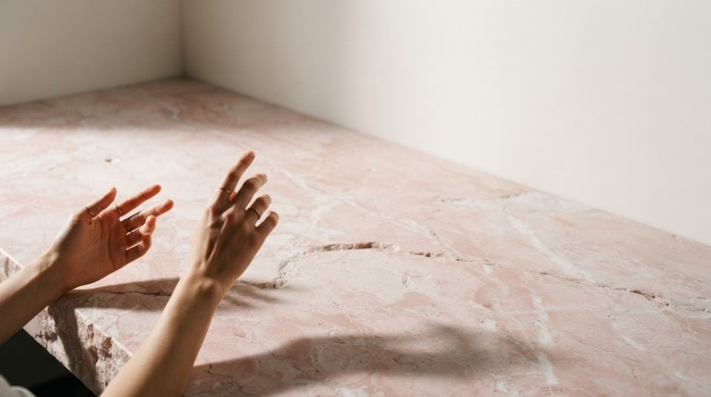Mãos femininas sobre superfície de mármore rosa com parede branca ao fundo