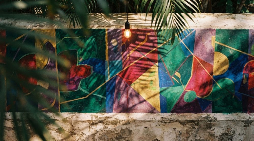 Mural colorido de arte abstrata numa parede exterior com sombras de plantas e luz natural