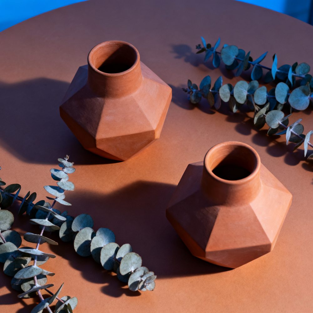Dois vasos geométricos em terracota com ramos de folhagem seca numa mesa laranja sob luz azul