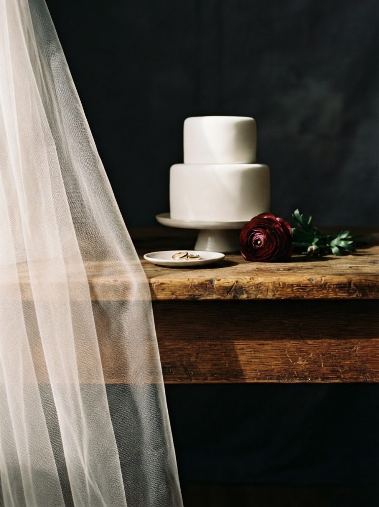 Bolo de casamento branco de dois andares com alianças num prato e flor vermelha sobre mesa de madeira