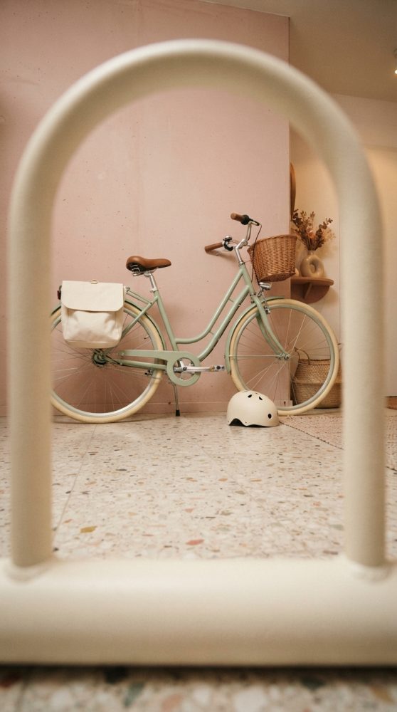 Bicicleta urbana verde pastel com cesta, saco branco e capacete em interior com parede rosa e piso de terrazzo