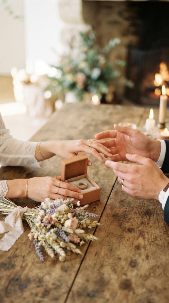 Mãos de casal trocando alianças sobre mesa de madeira com buquê rústico e velas acesas
