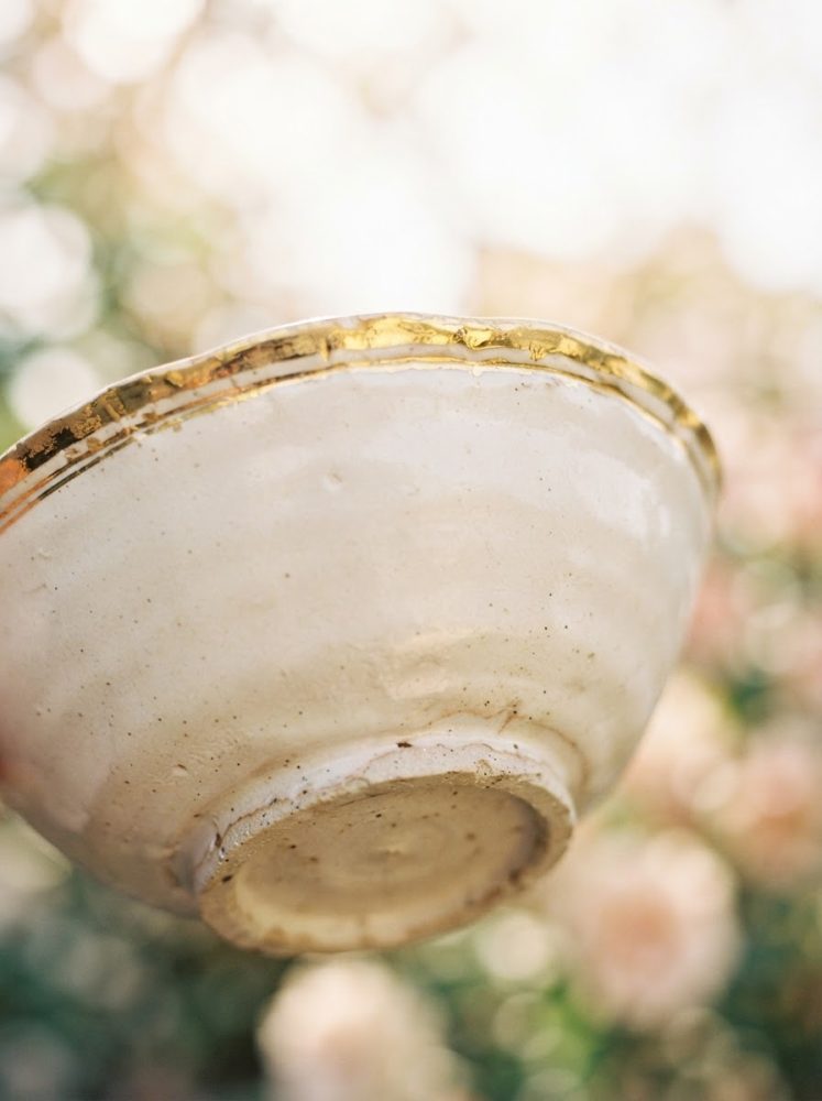 Taça artesanal de cerâmica com acabamento dourado sobre fundo desfocado de flores