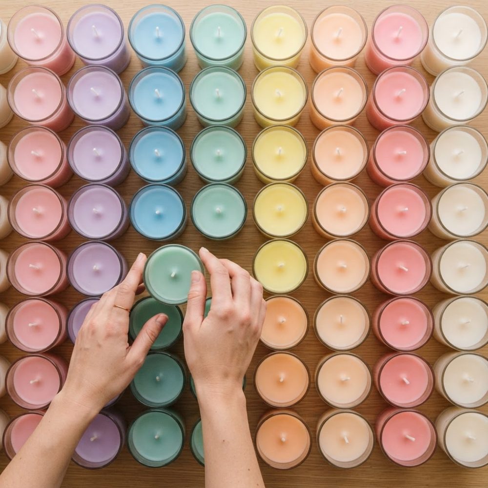 Mãos a segurar vela verde cercada por velas coloridas em copos sobre madeira