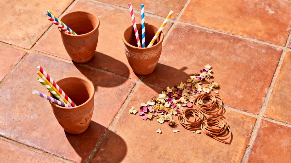 Copos de barro com canudos coloridos e confetes florais secos sobre chão de tijoleira