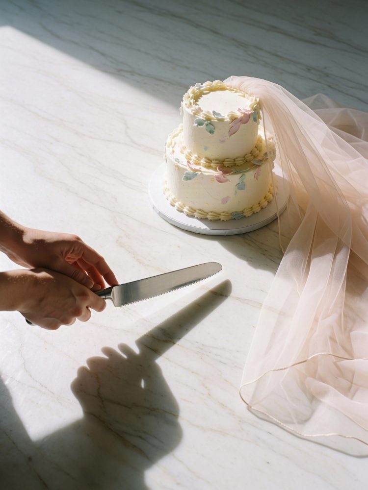 Bolo de casamento branco de dois andares com decoração floral e véu ao lado em mesa de mármore