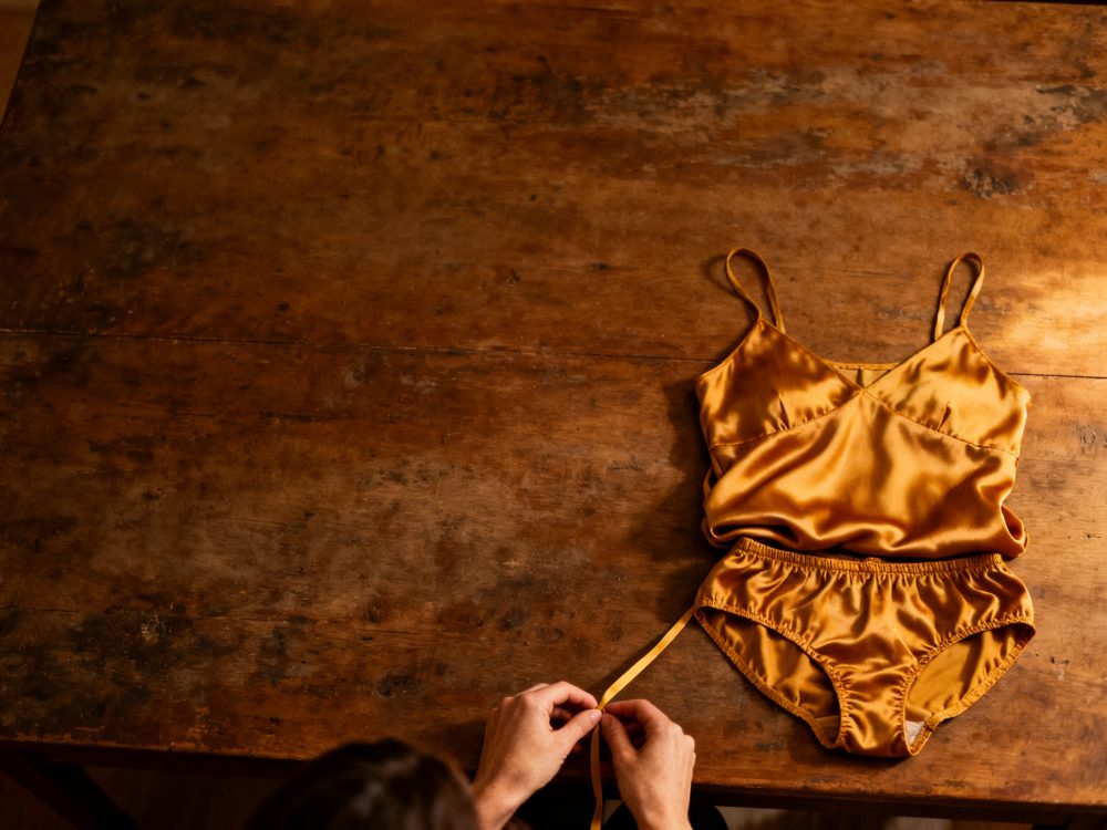 Conjunto de lingerie dourada em cetim sobre mesa de madeira com mão segurando fita métrica