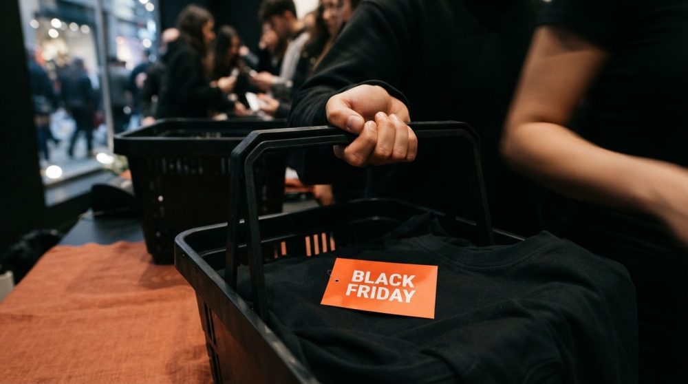 Mão segura cesta preta com roupa preta e etiqueta vermelha Black Friday numa loja com clientes desfocados