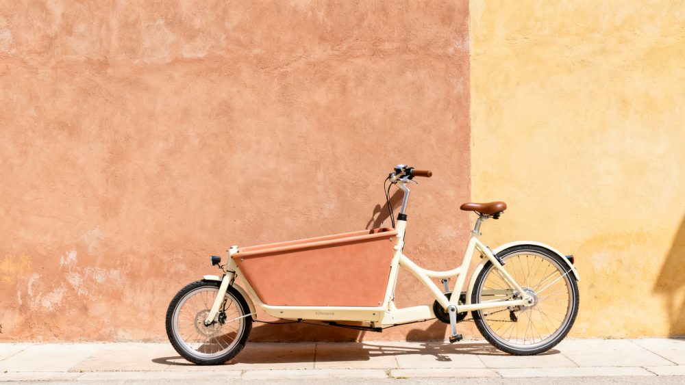 Bicicleta elétrica de carga bege com assento castanho contra parede texturada terracota e amarela