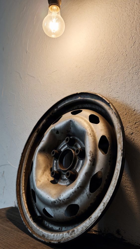 Jante metálica velha e oxidada apoiada contra parede iluminada por lâmpada incandescente.