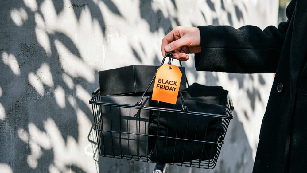Pessoa a segurar cesto de compras preto com etiqueta Black Friday e roupas negras contra parede cinzenta