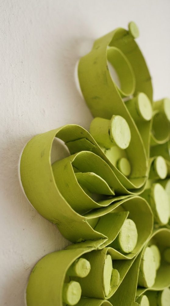 Escultura abstrata verde com formas curvas em relevo montada numa parede branca