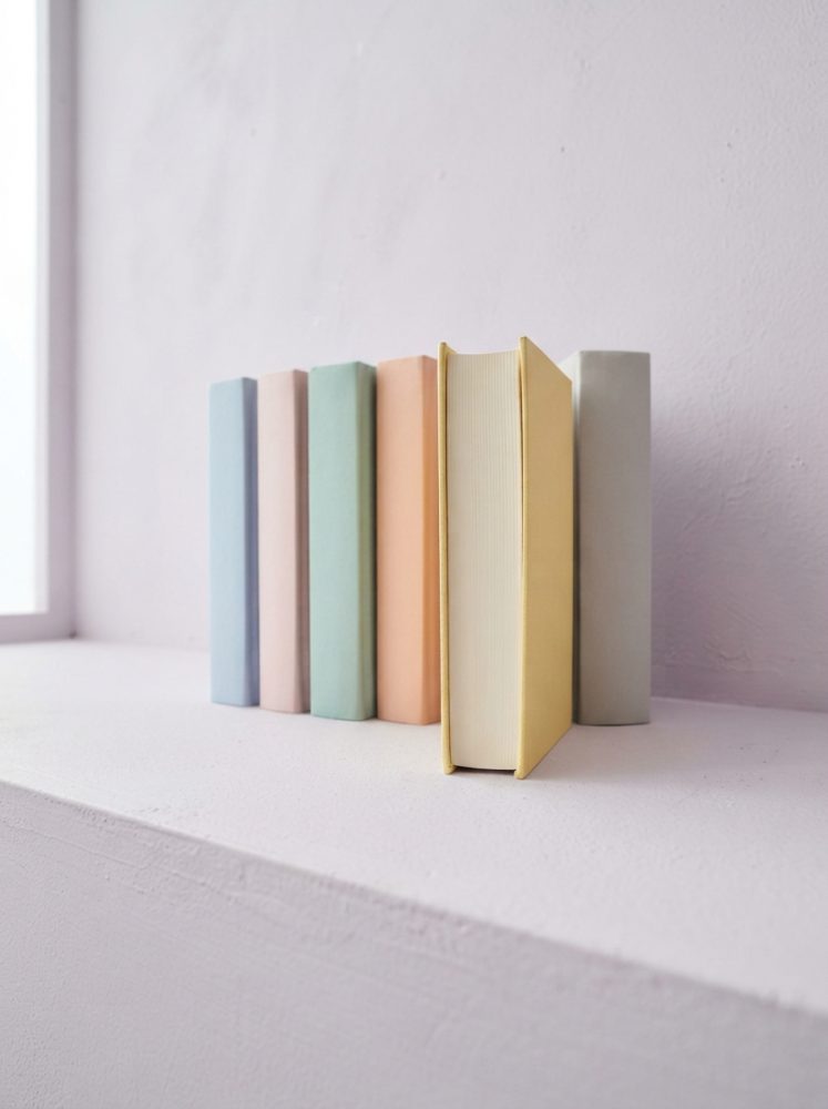 Seis livros com capas coloridas em tons pastel dispostos numa prateleira branca junto à parede clara.