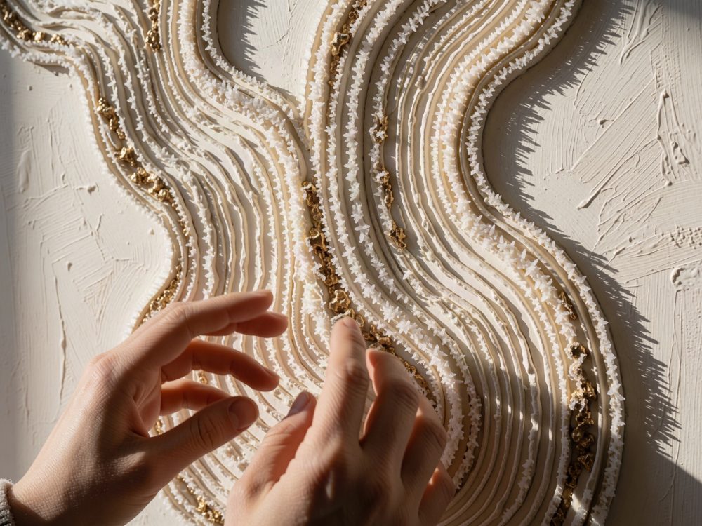 Mãos manipulando arte abstrata em camadas texturadas bege, branca e dourada com sombras.
