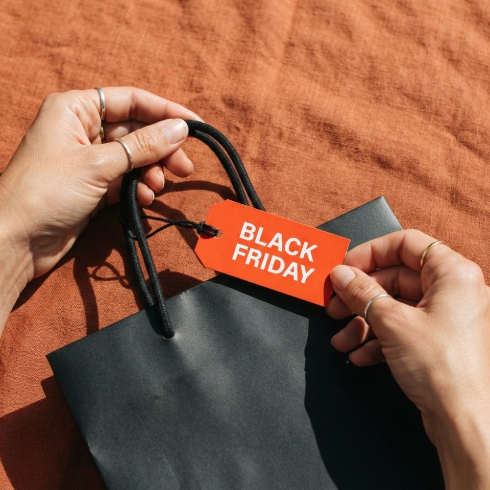 Mãos segurando etiqueta vermelha com Black Friday numa sacola preta sobre tecido castanho