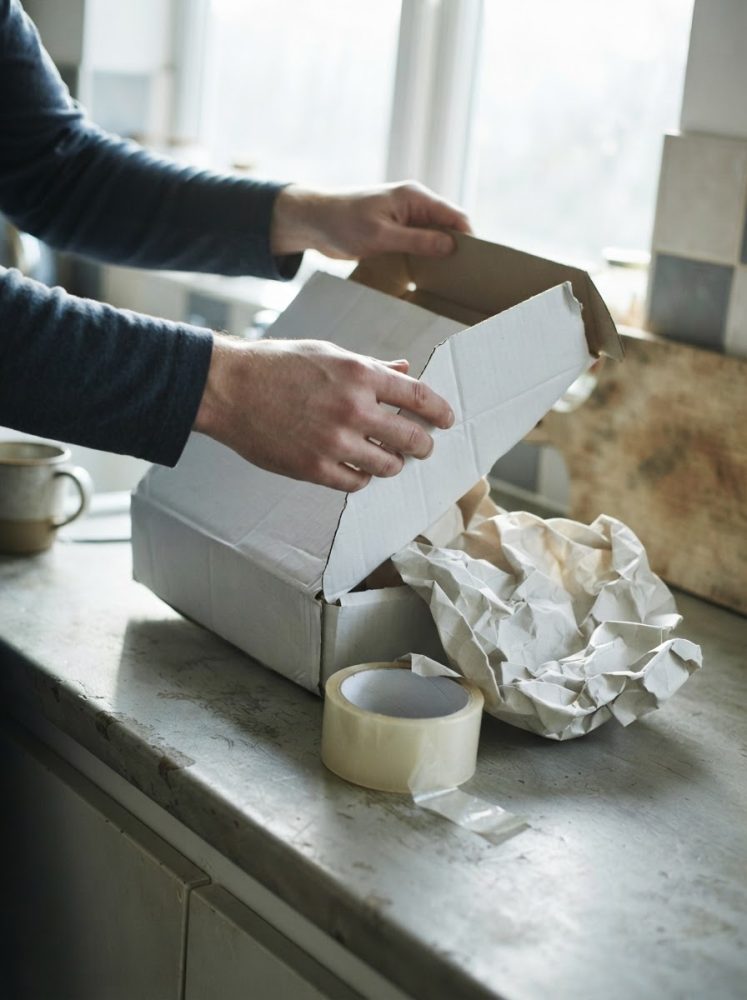 Mãos a fechar caixa de cartão com papel amassado e fita cola numa bancada clara