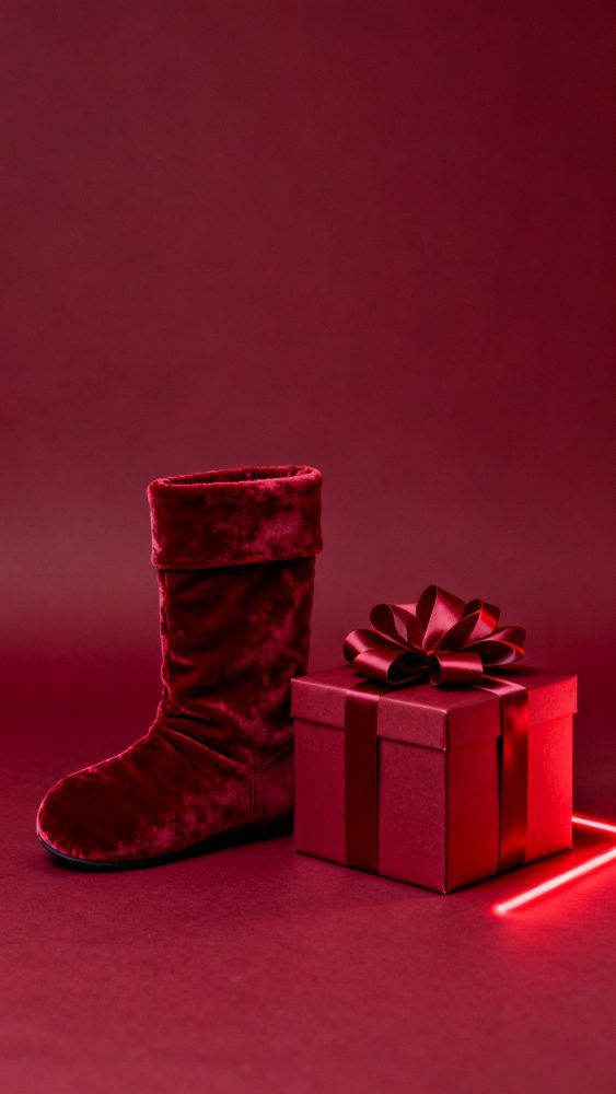 Bota vermelha de veludo ao lado de caixa de presente vermelha com laço, fundo vermelho