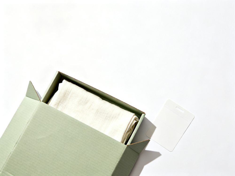 Caixa de papelão verde aberta com tecido branco dobrado e etiqueta branca ao lado