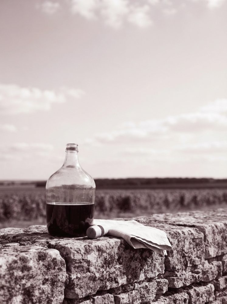 Garrafa de vinho tinto com rolha e pano branco sobre muro de pedra com vinhas ao fundo