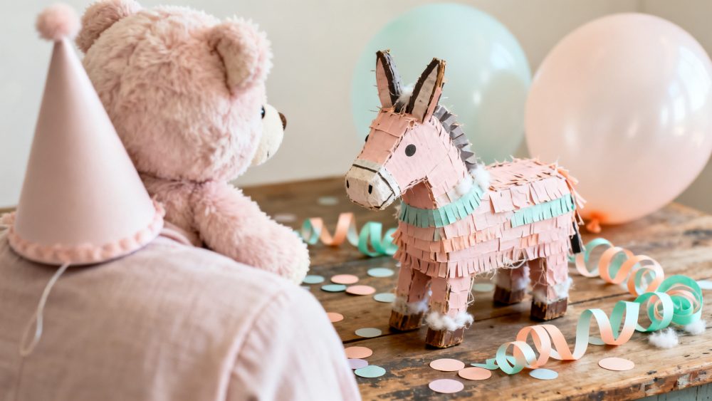 Piñata em forma de burro, ursinho rosa com chapéu de festa e balões pastel sobre mesa de madeira.