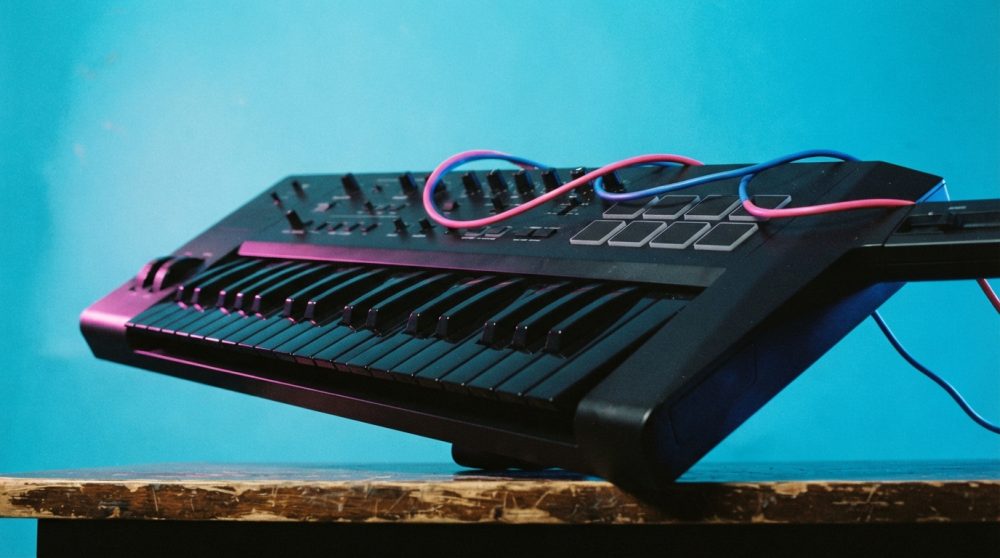 Teclado MIDI preto com cabos vermelho e azul sobre mesa de madeira rústica com fundo azul
