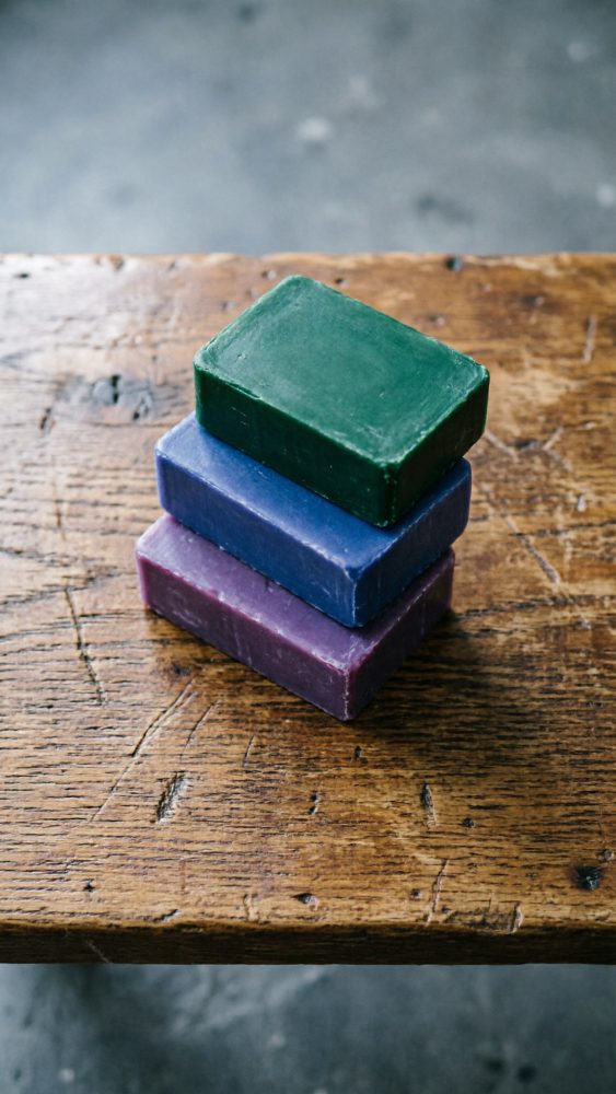 Três barras retangulares de sabão verde, azul e roxo empilhadas sobre mesa de madeira rústica