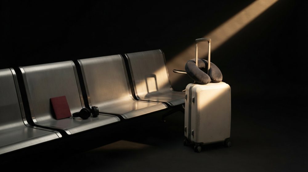 Mala branca com almofada de pescoço e acessórios num banco metálico sob luz focada em ambiente de aeroporto