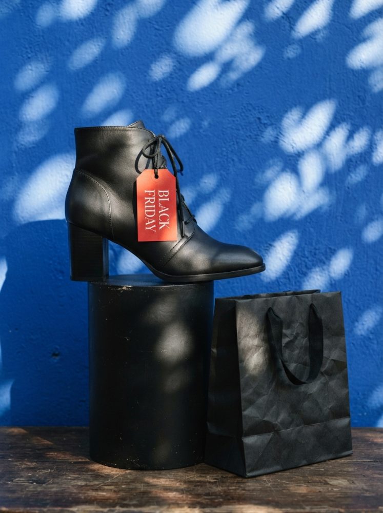 Bota preta de salto com etiqueta vermelha Black Friday ao lado de saco de compras preto sobre fundo azul