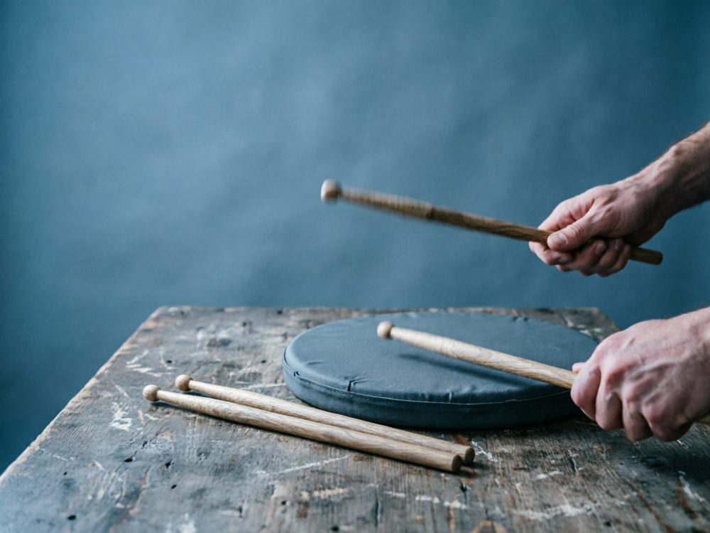 Mãos a tocar almofada de prática com baquetas de bateria sobre mesa de madeira rústica
