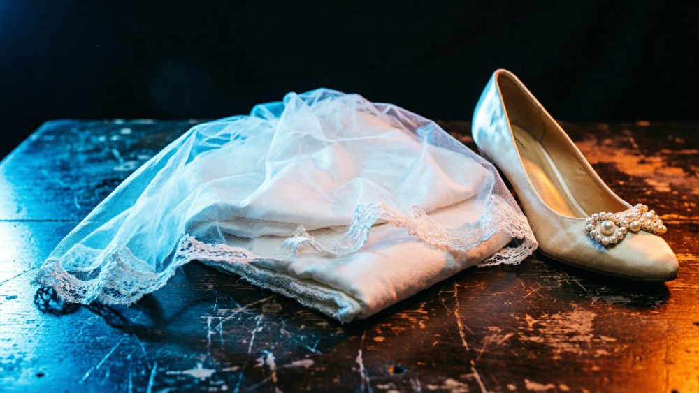 Véu rendado de noiva e sapato branco com pérolas sobre mesa de madeira escura