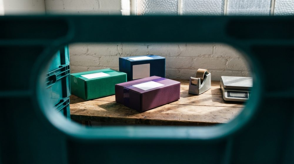 Caixas de envio coloridas numa mesa com fita adesiva e balança vista através de abertura azul
