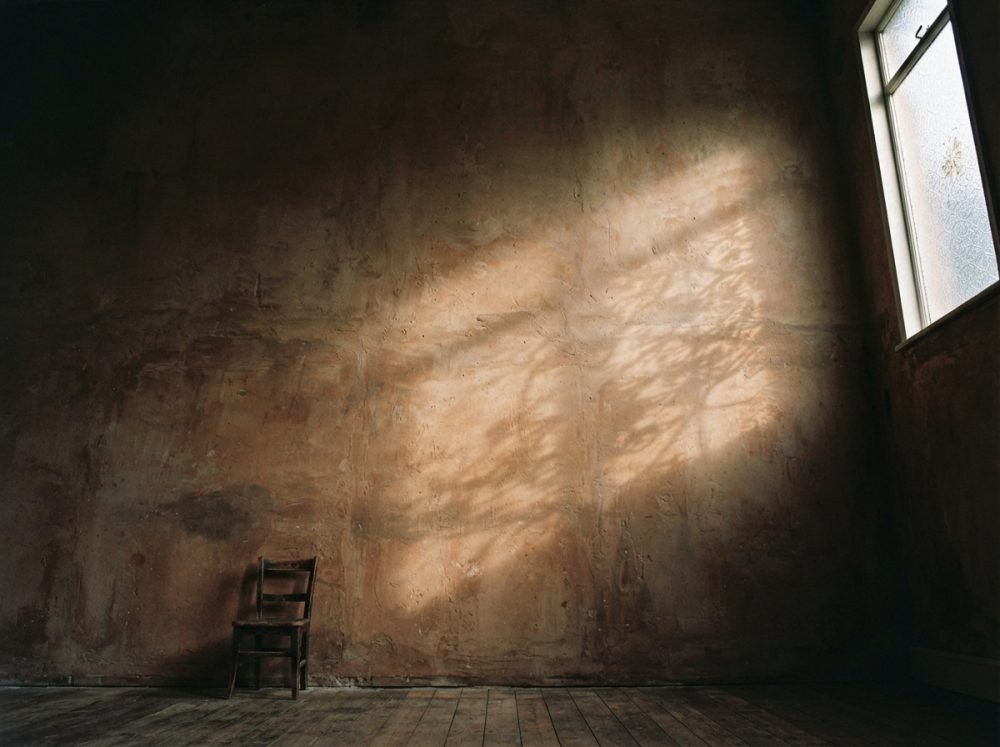 Parede rústica iluminada pela janela com sombras de folhas e cadeira de madeira junto ao chão