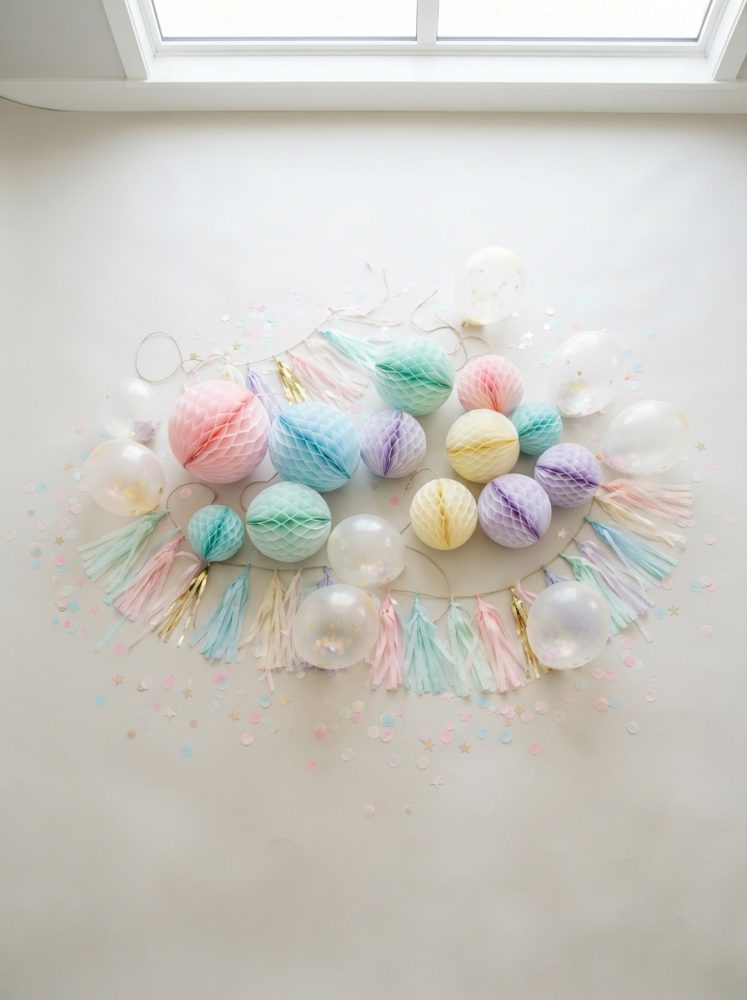 Decoração de festa com bolas de papel em tons pastel, balões transparentes com confetis e franjas coloridas