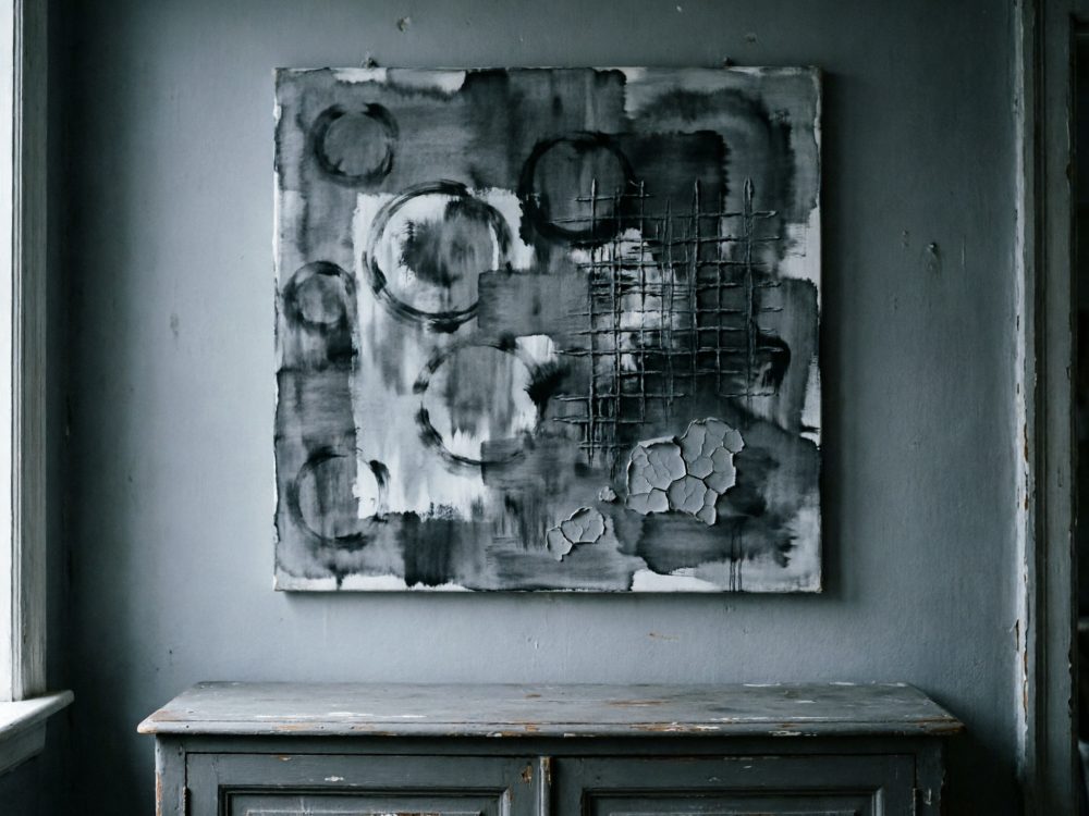 Quadro abstrato a preto e branco com círculos e linhas texturizadas numa parede cinzenta acima de móvel antigo de madeira