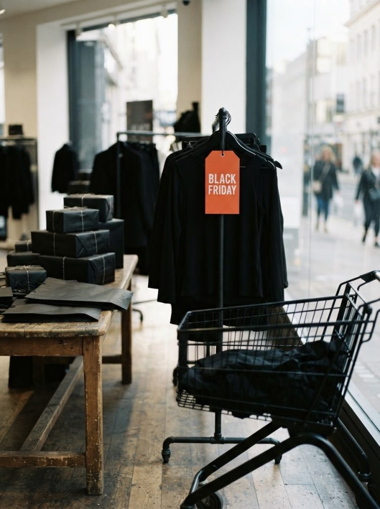 Roupa preta com etiqueta Black Friday pendurada numa loja urbana com carrinho de compras e mesa de madeira