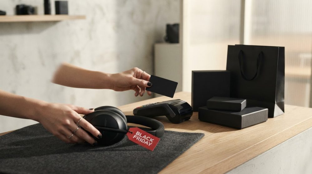 Mãos seguram fones de ouvido com etiqueta Black Friday e cartão perto de embalagens e máquina de pagamento