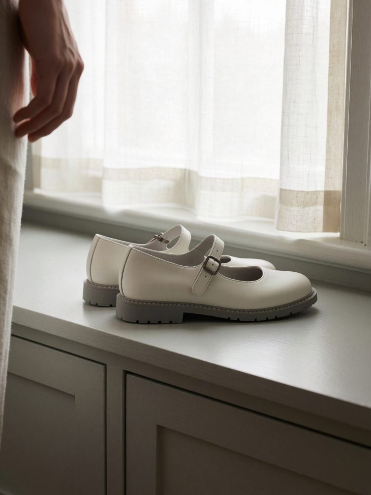 Sapatos brancos tipo Mary Jane com sola cinza sobre móvel branco junto a janela com cortinas