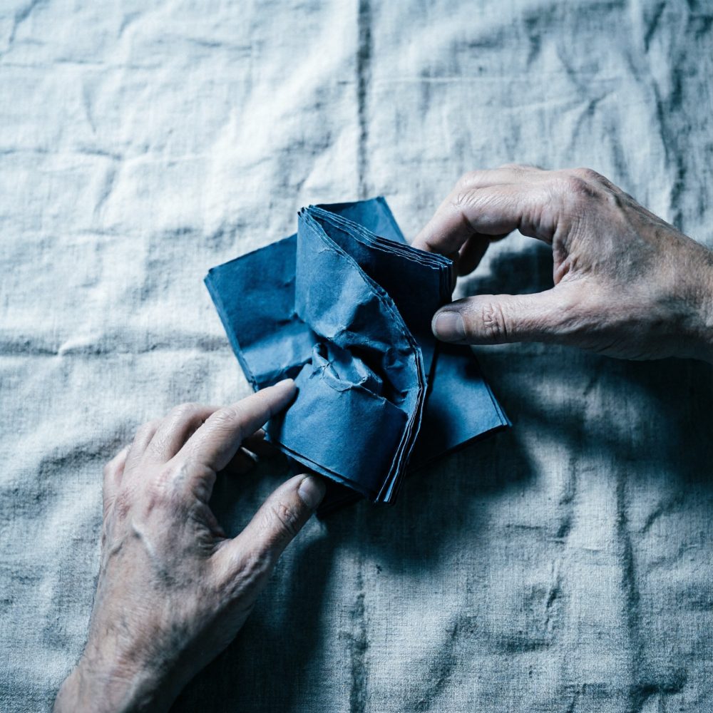 Duas mãos manipulam papel azul amassado sobre tecido cinzento claro texturado