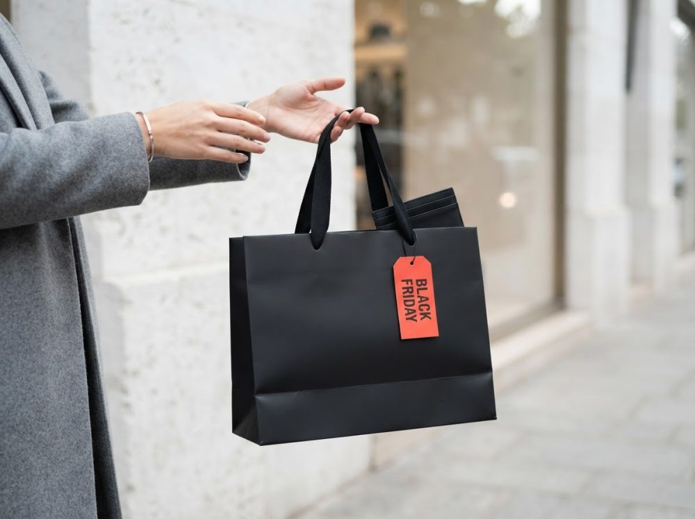 Mãos a segurar saco de compras preto com etiqueta vermelha Black Friday numa rua citadina.