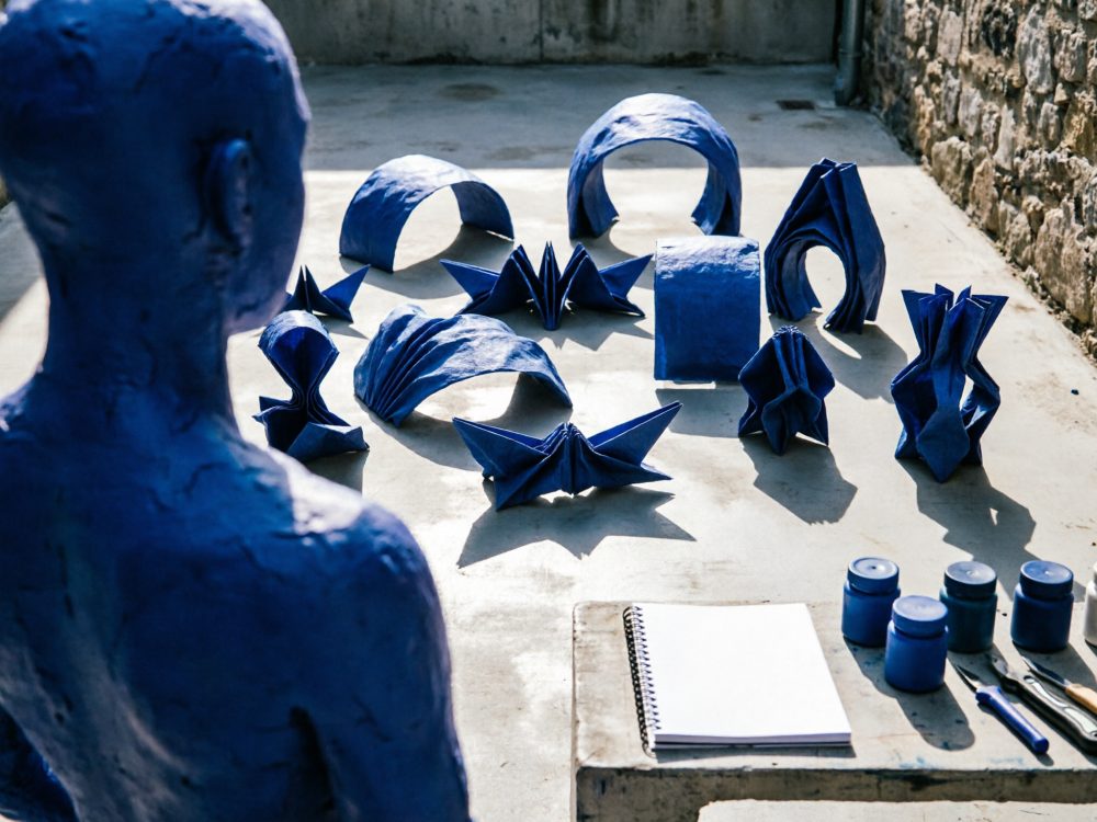 Escultura humana azul voltada para várias esculturas abstratas também azuis sobre chão de cimento em espaço rústico