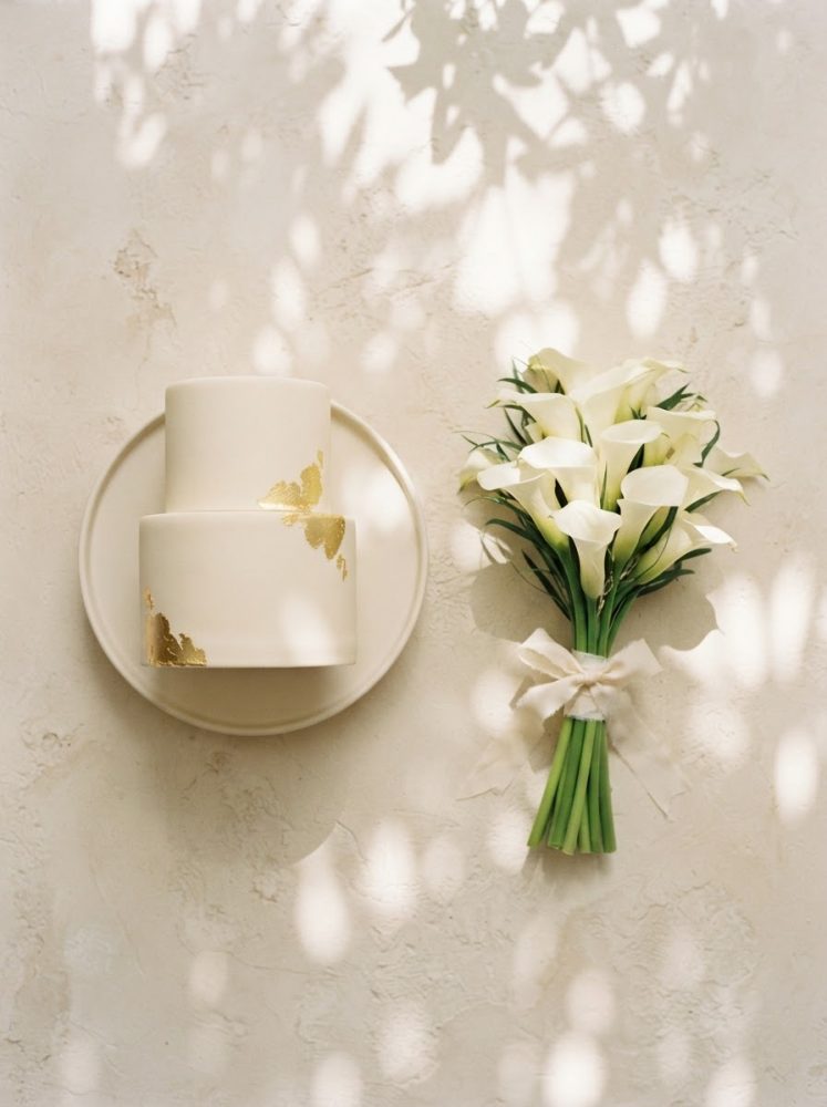 Bolo branco de casamento com detalhes dourados e bouquet de copos-de-leite brancos