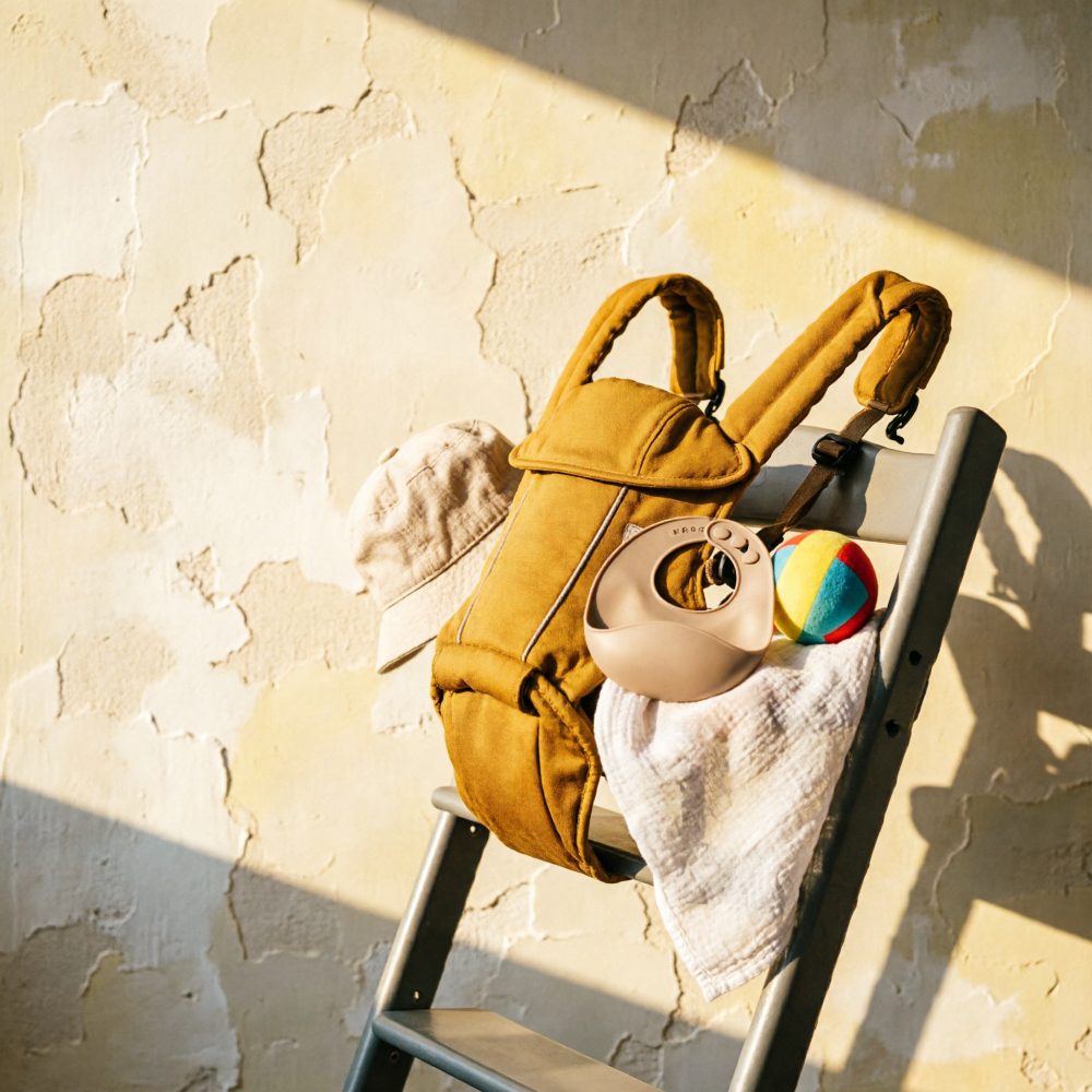 Mochila castanha, chapéu bege, mantinha branca e bola colorida numa cadeira cinzenta com luz solar