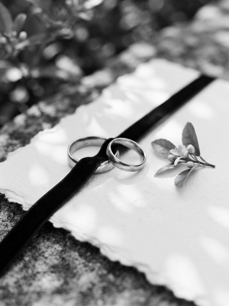 Alianças de casamento sobre papel branco com fita preta e flor branca ao lado