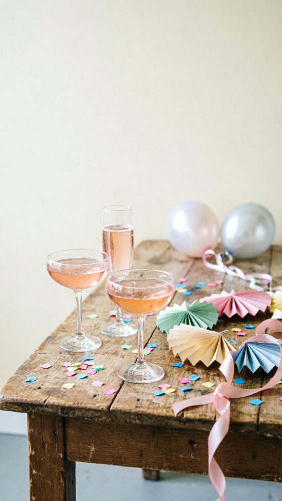 Mesa de madeira com copos de cocktail rosé, confetis coloridos, decorações em papel e balões festivos