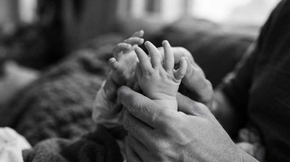 Mãos de adulto segurando ternamente as mãos de um bebé em preto e branco