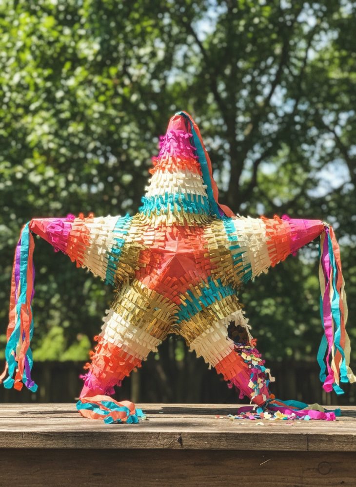 Piñata em forma de estrela colorida com franjas penduradas, sobre mesa de madeira, fundo com árvores verdes desfocadas