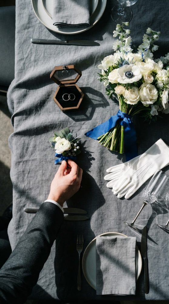 Mesa de casamento com bouquet, alianças em caixa de madeira, luvas brancas e mão a ajustar flor