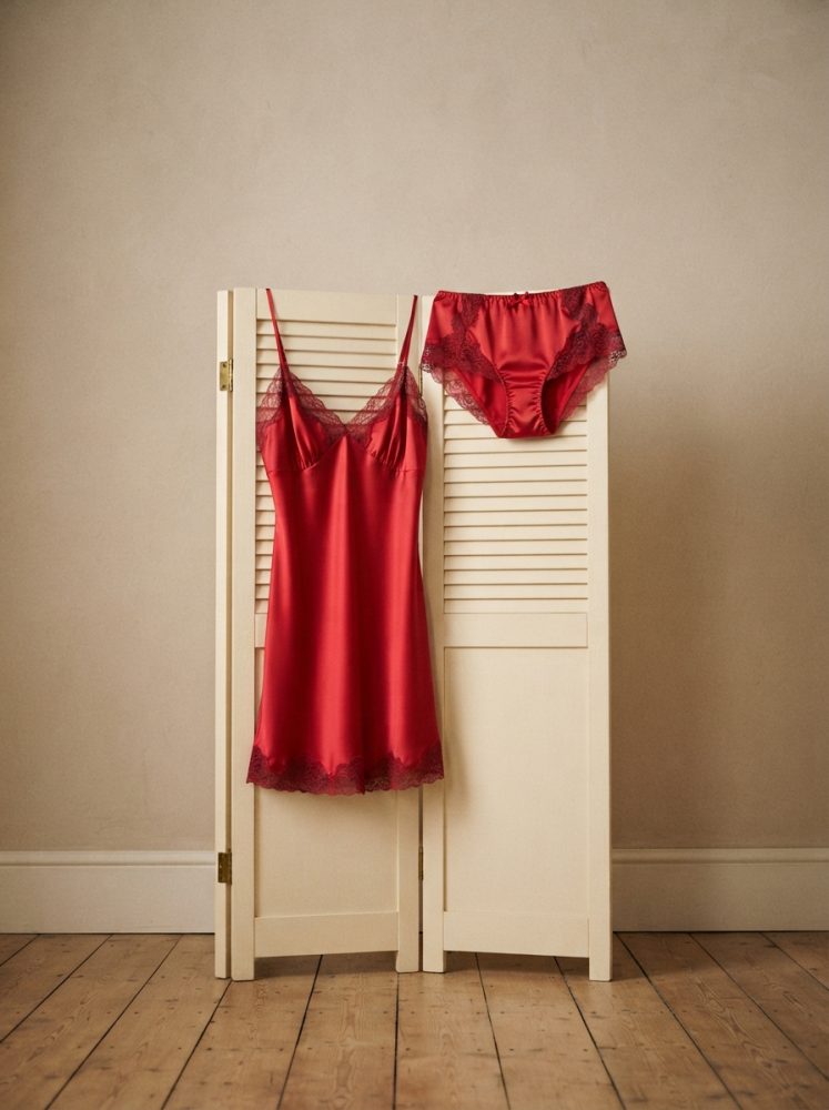 Conjunto vermelho de lingerie em cetim com renda pendurado num biombo branco