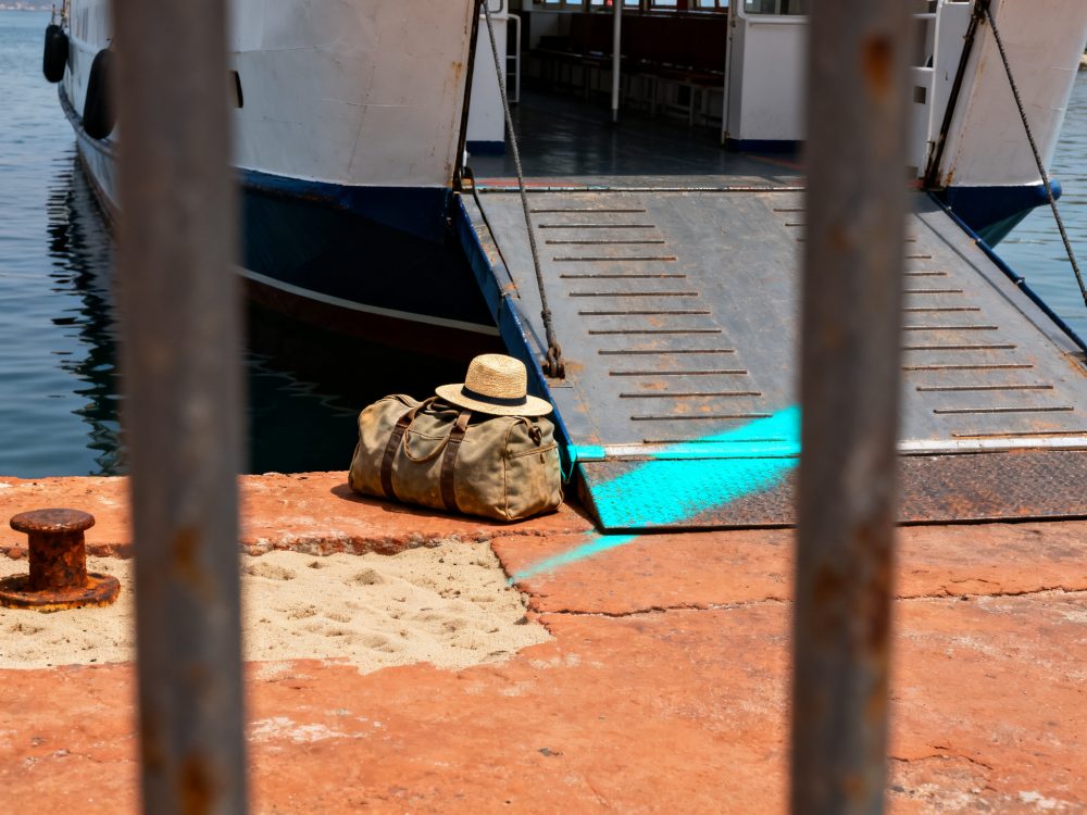 Mala de viagem com chapéu de palha no cais ao lado da rampa de ferry ancorado em porto