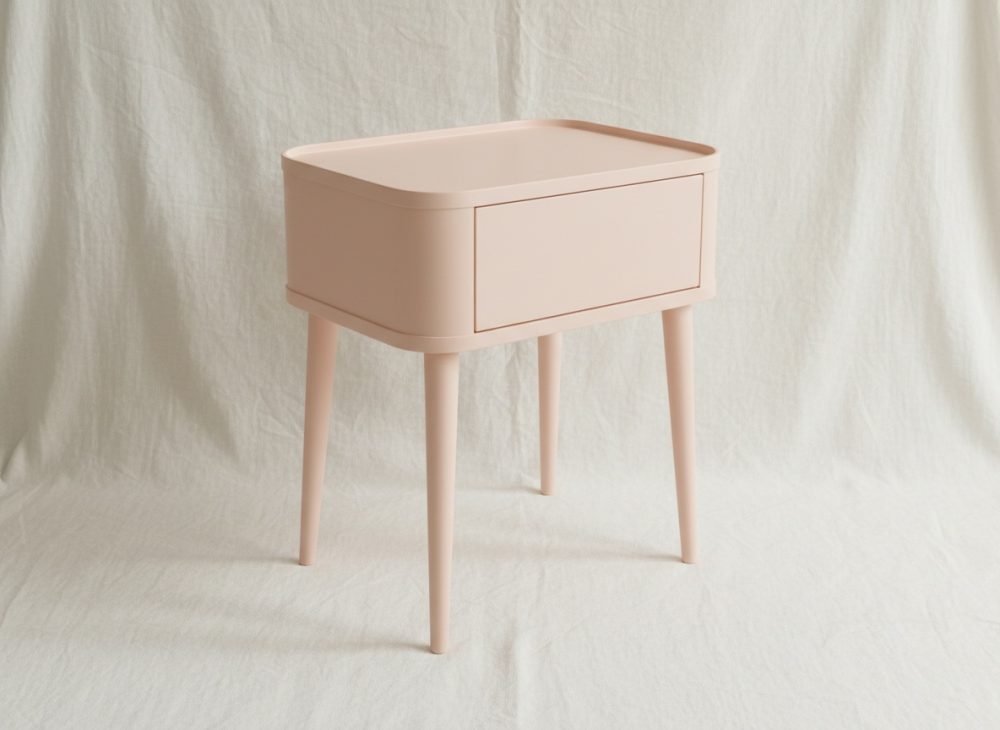 Mesa de cabeceira rosa claro com gaveta, pernas esguias e fundo neutro